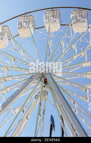 Low-angle shot of a modern white Ferris wheel structure with cabins against a vibrant clear blue sky. Stock Photo