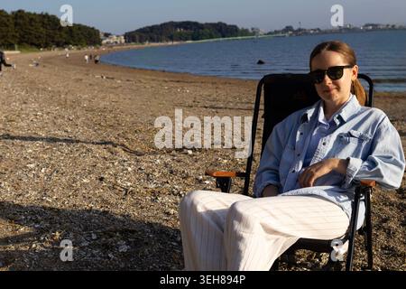 Young woman wearing sunglasses relaxing on a beach chair near the sea during golden hour. Summer vacation, travel and coastal lifestyle concept Stock Photo