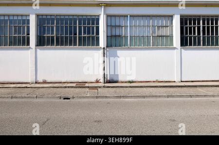 White wall with large windows of an old industrial building. Sidewalk and asphalt road in front. Background and texture Stock Photo