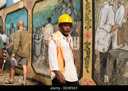 Sri Lanka - 20 February 2026: View of a worker wearing a yellow hard hat and orange safety vest walking past a wall decorated with colorful murals. Stock Photo