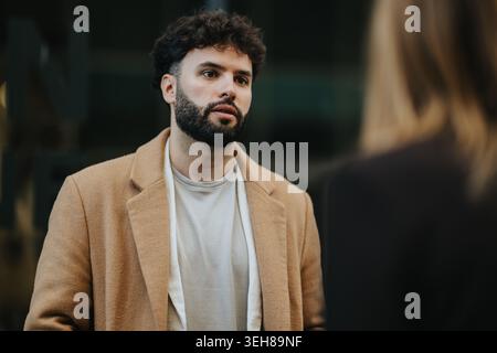 Young bearded man in tan coat engaged in serious conversation on an urban street Stock Photo