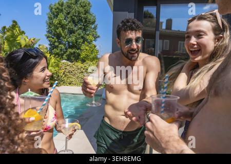 Happy multiracial friends laughing and clinking cocktails poolside at a sunny villa summer party, enjoying drinks, conversation and relaxed vacation v Stock Photo