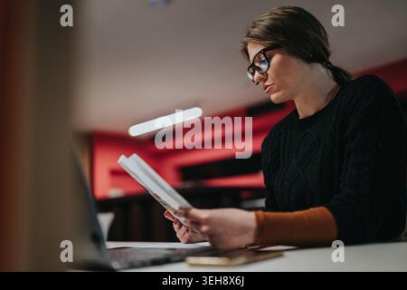 Businesswoman reading documents at desk with laptop in modern office workspace, focused and professional Stock Photo