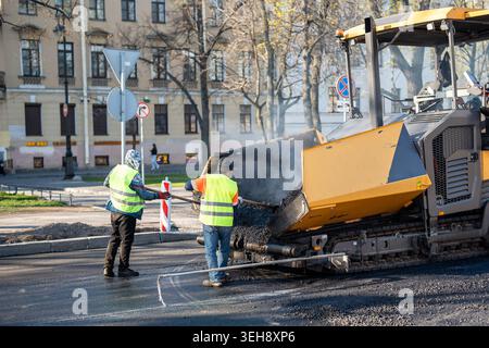 Road workers in vests using shovels tools to scoop fresh hot asphalt from paver machine onto road. Stock Photo