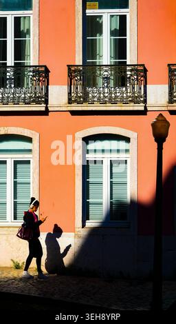 Woman on smartphone cast in dramatic shadow against vivid orange facade, Alcântara, Lisbon, Portugal Stock Photo