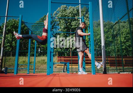 Two young athletes doing outdoor calisthenics workout on blue pull-up bars in a sunny park Stock Photo