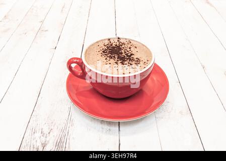 A red coffee cup with creamy froth and cocoa powder sits centered on a matching saucer over whitewashed wooden planks. Stock Photo