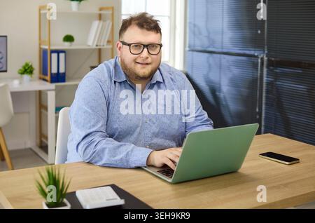 Overweight freelancer man working on laptop at modern home office desk, smiling Stock Photo
