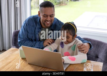 Father and daughter sitting at home, father leaning while girl typing on laptop with mug, headband Stock Photo