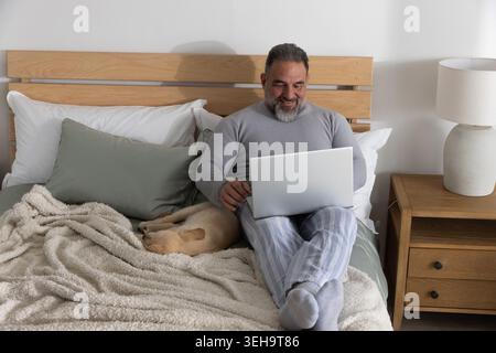 Mature man sitting on bed in neutral bedroom, using silver laptop, small tan dog sleeping Stock Photo