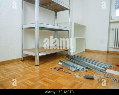 Metal shelving unit is partially assembled in a room with a wooden floor, tools and materials scattered on the ground, and an empty white shelf nearby Stock Photo