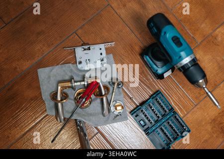 Tools and hardware for door lock installation or repair project, including disassembled handle, latch mechanism, power drill, and screwdriver, laid Stock Photo