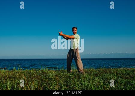 Man in sportswear standing on grass with sea, mountains and sky in background, making gun shape with hands Stock Photo