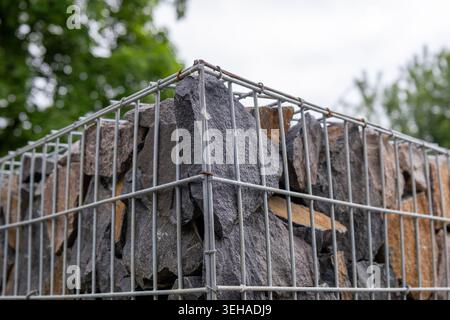 A structure made of stone is surrounded by a metal cage. The stones are stacked neatly. The scene shows trees and a cloudy sky in the background. Stock Photo