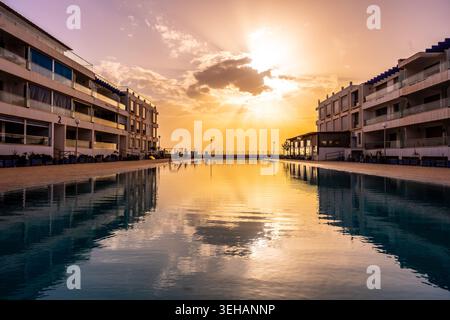 People enjoy the sunset by the pool at a summer resort. The sun sets behind buildings, creating reflections on the water. It is a warm evening, perfec Stock Photo