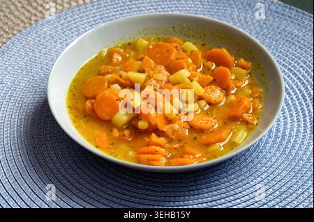 A fresh, healthy bowl of traditional vegetable stew with sliced carrots and diced potatoes, served on a blue textured placemat in daylight Stock Photo