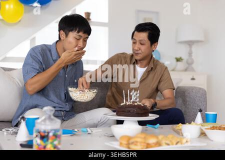 Asian male friends sitting on couch at home sharing chocolate cake with popcorn bowl Stock Photo