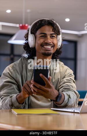 Indian man sitting at modern cafe smiling while holding smartphone wearing white headphones Stock Photo