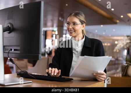 Woman receptionist holding documents, working on computer at reception desk Stock Photo
