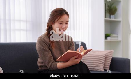 Young Asian woman writing in notebook while sitting on couch at home taking notes planning organizing work Stock Photo