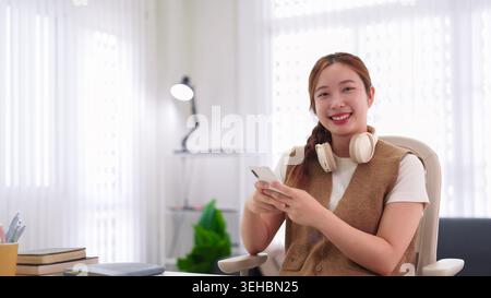 young Asian businesswoman using smartphone with headphones at home office desk smiling happy relaxed casual workspace Stock Photo