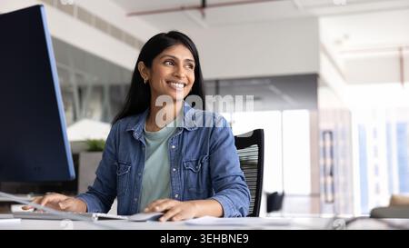 Cheerful Indian businesswoman sits at desk working on pc Stock Photo