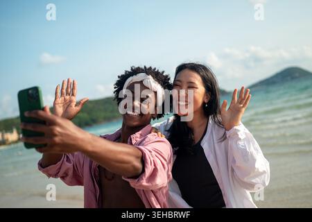 Diverse couple connected, embracing technology and social media while enjoying moments at the beach Stock Photo
