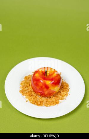 Glazed red mousse cake shaped like an apple served on a plate with crispy flakes under a large green space Stock Photo