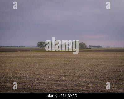 Distant rural white farmhouse and green trees nestled among vast agricultural fields under a dramatic overcast stormy sky Stock Photo