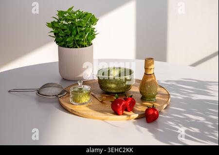 Stylish arrangement of matcha tea accessories displayed with soft studio lighting on clean surface with potted plant. Ceramic bowl and sieve with past Stock Photo