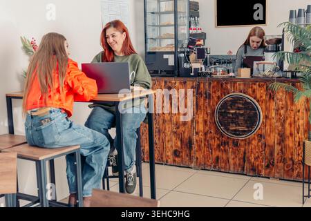 mother and daughter sit in a cafe. The mother has red hair and wears a dark sweater. The daughter has long brown hair and wears an orange top. They Stock Photo