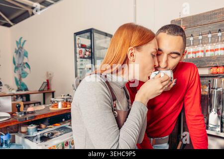 red-haired woman and a young man share a moment at a cafe. The woman sips coffee while the man leans in, smiling. The cafe has a modern decor. Stock Photo