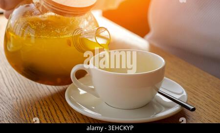 Glass Teapot Pouring Hot Lemon Tea, White Ceramic Cup Catching Amber Pour On Sunlit Wooden Table Soft Steam Rising, Spoon Resting On Saucer, Cozy Stock Photo