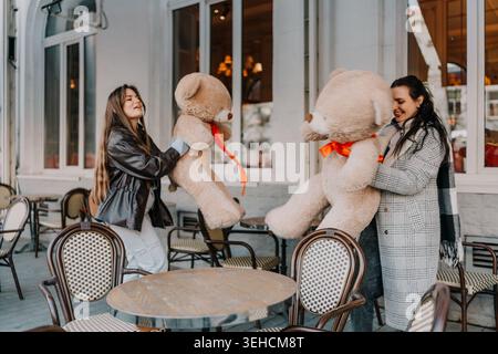 Friends teddy cafe outdoor terrace where two young women laugh while playing with large plush bears on a sunny afternoon Stock Photo