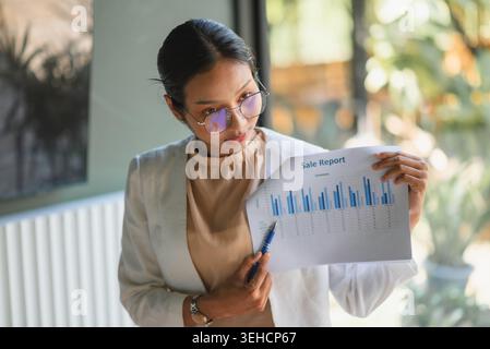 Businesswoman pointing to sales report with pen in bright office, analyzing bar chart data for finance review, marketing strategy, business presentati Stock Photo