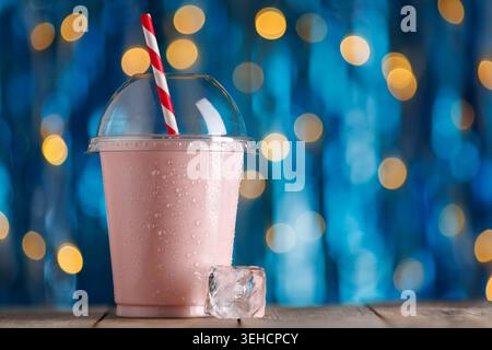 Strawberry milkshake in disposable plastic cup with condensation and red and white striped straw on wooden surface Stock Photo