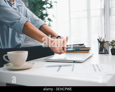 Professional stretching hands after closing laptop on white office desk with coffee cup, notebooks, and reports, showing work break, wellness, product Stock Photo