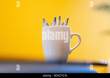 A white mug full of pens sits on a table in front of a yellow background Stock Photo