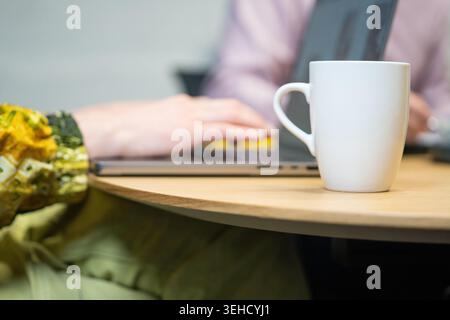 White mug on a desk while a woman types on a laptop in the background Stock Photo