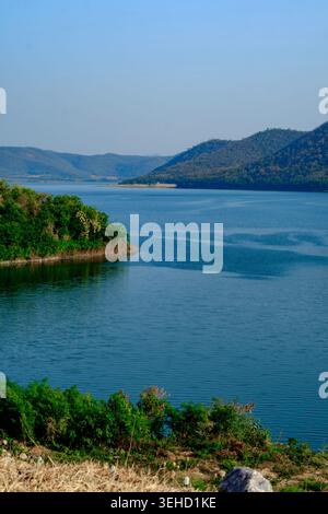 Peaceful panoramic view of a deep blue lake nestled between rolling hills and green vegetation under a clear bright sky. Stock Photo
