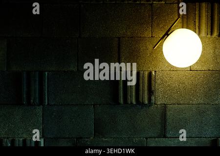 A minimalist spherical lamp casts a warm glow across a dark industrial cinder block wall, featuring ample copy space and shadows Stock Photo