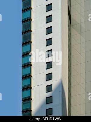 A clean, vertical architectural detail of a white skyscraper featuring blue-tinted windows, geometric patterns, and sharp shadows against a clear sky Stock Photo