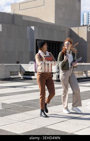 Diverse female students walking across plaza by concrete building wearing beanie carrying notebooks. Learners, campus, outdoor, urban, architecture, backpack, folders Stock Photo
