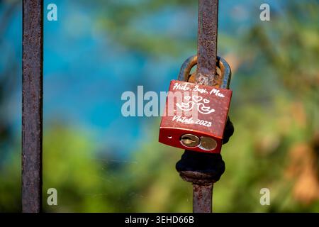 Vieste - Apulia - Italy - 08-29-2025: Red padlock with handwritten names and hearts hangs on a rusty iron fence, with blurred green and blue background in bright daylight. Stock Photo