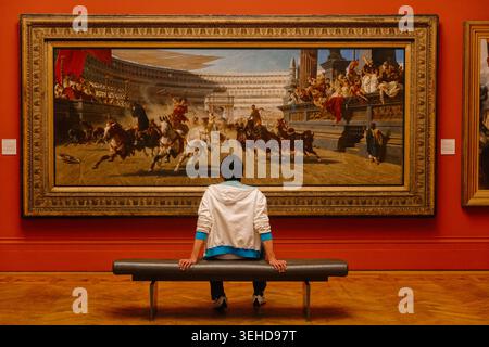 A visitor sitting on a bench looks at Alexander von Wagner's monumental painting The Chariot Race at Manchester Art Gallery, England, UK Stock Photo