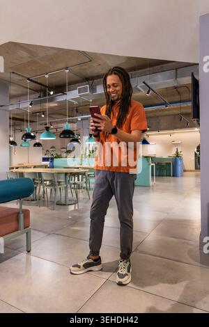 African American man in 20s wearing orange tee standing in coworking space holding phone smiling Stock Photo