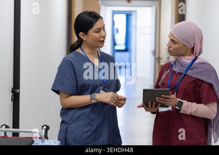 Two female medical workers in scrubs standing in hospital corridor discussing tablet, stethoscope Stock Photo