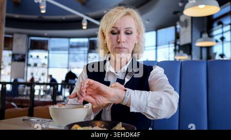 Elderly Businesswoman Checking Watch During Lunch In Modern Cafe Booth, Stirring Soup While Glancing At Wristwatch, Wearing Blazer And White Blouse Stock Photo