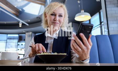 Caucasian Elderly Businesswoman Checking Calendar On Smartphone During Lunch At Modern Cafe, Concentrated Expression, Fork At Plate, Digital Stock Photo