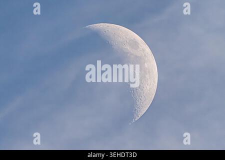 Detailed crescent moon shining through thin clouds in bright daytime sky. Atmospheric lunar photography featuring visible craters, soft blue tones and Stock Photo